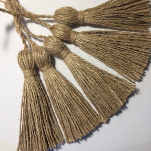 May include: Close-up of several natural jute tassels. Each tassel features a rounded top and a long, fringed bottom. The tassels are a warm, earthy brown colour, and are arranged against a white background. These tassels are ideal for craft projects.