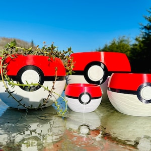 May include: Four decorative planters in the shape of a Poké Ball, in red, white, and black. The largest planter contains a green plant. The planters are of varying sizes, with a smooth, glossy finish. The background is a clear blue sky.