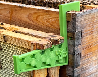 May include: Green plastic beehive frame spacer with a honeycomb pattern and a bee graphic. The spacer is attached to a wooden beehive.