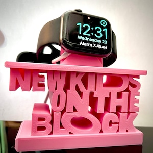 May include: Pink 3D printed phone stand with the words "New Kids on the Block" in white letters. An Apple Watch is charging on the stand.