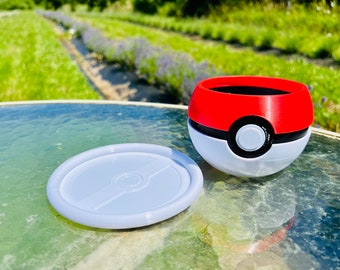 May include: A red, white, and black Pokeball-shaped container with a lid. The lid is a flat, white disc with a black line across the center. The container is sitting on a glass table.