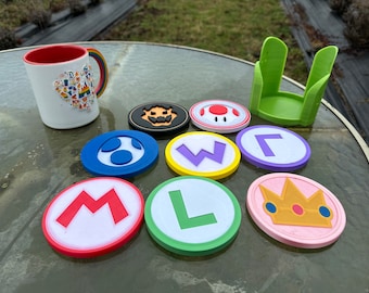 May include: A white mug with a rainbow handle and colorful icons, eight circular coasters featuring various character symbols, and a light green plastic coaster holder. The coasters display designs including the letters M, L, W, and L, a crown, a mushroom, a ghost, and a toadstool.