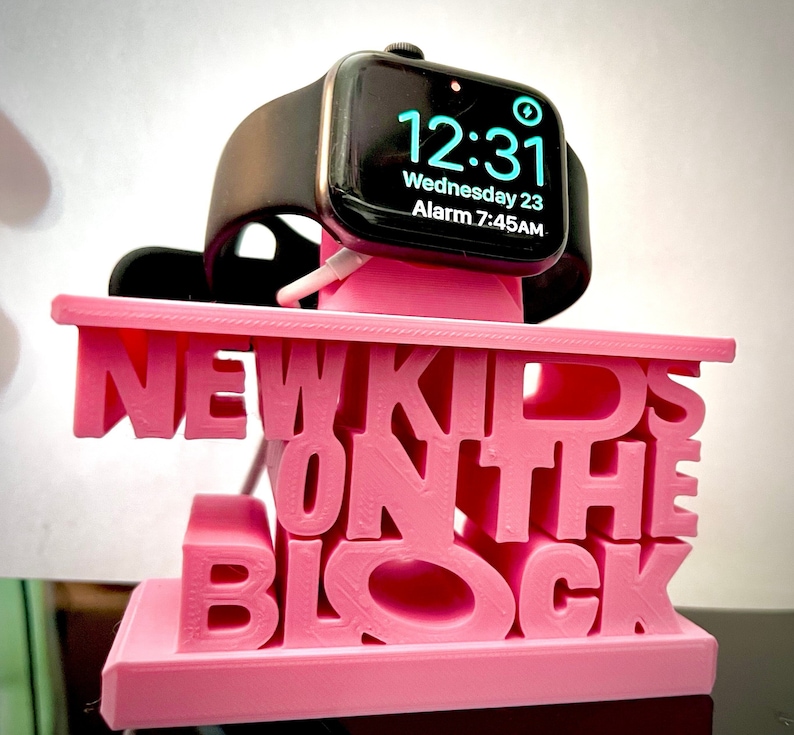 May include: Pink 3D printed phone stand with the words "New Kids on the Block" in white letters. An Apple Watch is charging on the stand.