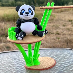 Peut inclure: Une figurine de panda noir et blanc assise sur une plateforme en bois brun et en bambou vert. La plateforme est soutenue par plusieurs tiges de bambou vert.