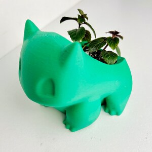 May include: A green 3D printed planter shaped like a Pokemon character, with a small plant growing inside.