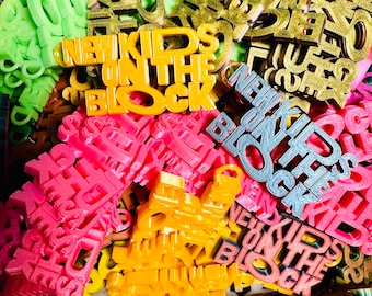 May include: A pile of colorful plastic charms with the words "New Kids on the Block" in various colors and styles. The charms are made of 3D printed plastic and have a glossy finish.