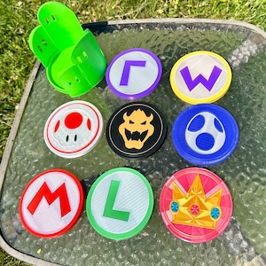 May include: Eight round, white coasters with different colored borders and designs. The designs include a red mushroom, a black Bowser, a blue star, a purple L, a green W, a pink crown, a red M, and a green L.