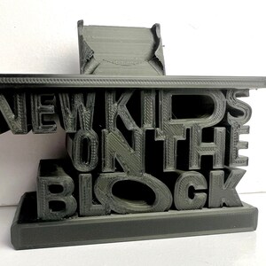 May include: A 3D printed black phone stand with the words "New Kids on the Block" in raised letters.