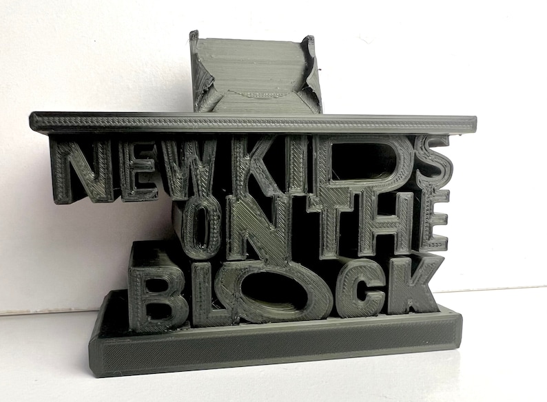 May include: A 3D printed black phone stand with the words "New Kids on the Block" in raised letters.