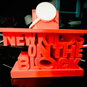 May include: A red 3D printed phone stand with the words "NEW KIDS ON THE BLOCK" in white letters. The stand has a small white circle on top for holding a phone.
