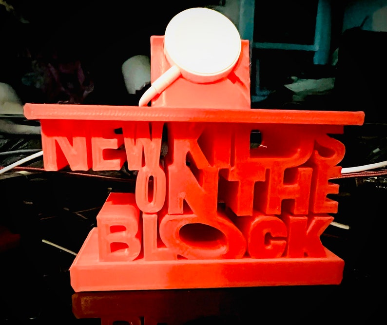 May include: A red 3D printed phone stand with the words "NEW KIDS ON THE BLOCK" in white letters. The stand has a small white circle on top for holding a phone.