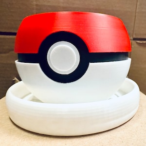 May include: A white and red 3D printed planter shaped like a Pokeball, with a black ring around the middle and a white circle in the center. The planter is sitting on a white ring-shaped base.
