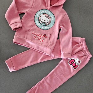 K&ouml;nnte beinhalten: Rosa Hello Kitty Kapuzenpullover und Jogginghosen-Set. Der Kapuzenpullover hat eine Vordertasche und eine runde Grafik mit Hello Kitty und dem Text "Just E You". Die Jogginghose hat einen elastischen Bund und eine Hello Kitty Grafik.