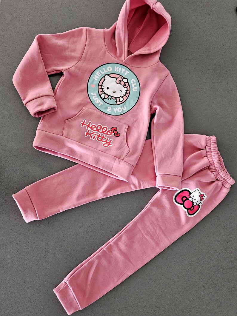 K&ouml;nnte beinhalten: Rosa Hello Kitty Kapuzenpullover und Jogginghosen-Set. Der Kapuzenpullover hat eine Vordertasche und eine runde Grafik mit Hello Kitty und dem Text "Just E You". Die Jogginghose hat einen elastischen Bund und eine Hello Kitty Grafik.