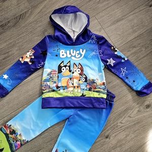 May include: A blue children's hoodie and matching pants set. The hoodie features the word "Bluey" and a graphic of cartoon dogs. The sleeves and pants have star and character graphics. The set is made of a soft material.