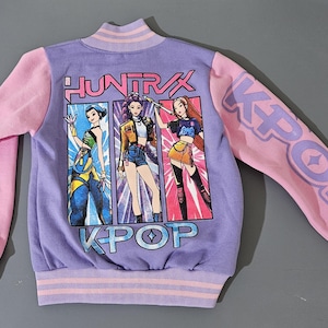Może przedstawiać: Liliowo-różowa kurtka bomberka KPOP. Kurtka posiada grafikę przedstawiającą trzy stylizowane postacie oraz napisy "HUNTRAX" i "KPOP". Rękawy są różowe z nadrukiem słowa "KPOP".