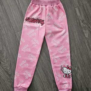 Op de afbeelding: Roze joggingbroek met een elastische tailleband en manchetten. De broek heeft een herhalend wit "Hello Kitty" patroon en een grote "Hello Kitty" afbeelding op de voorkant. Een cartoonafbeelding van Hello Kitty staat op het rechterbeen.