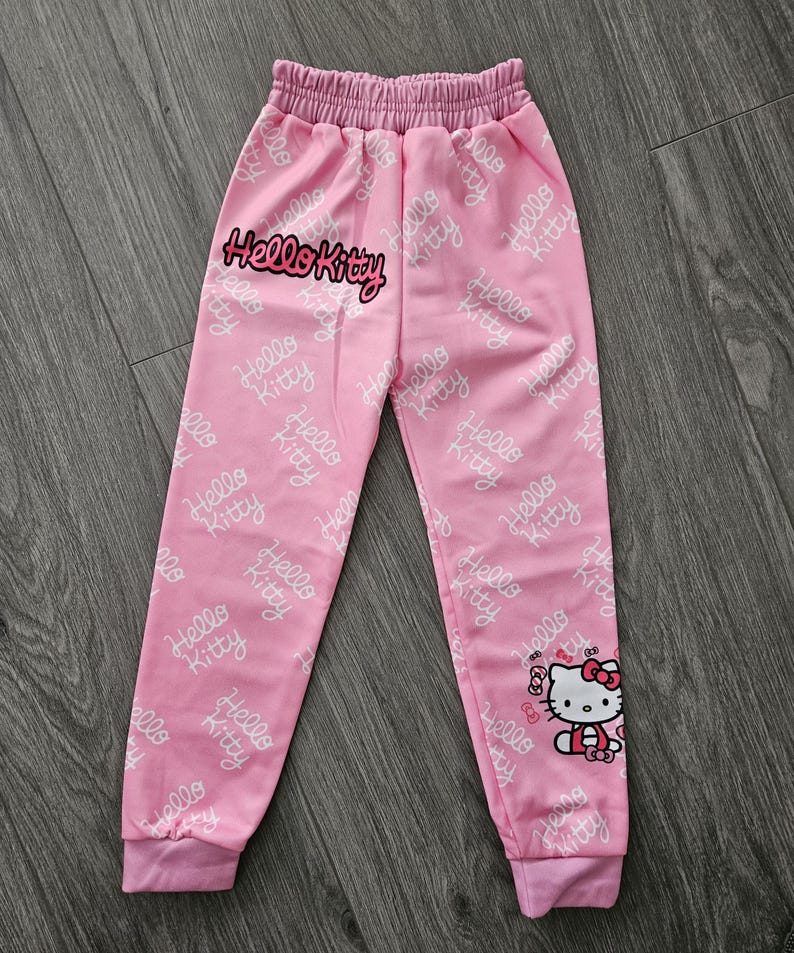 Op de afbeelding: Roze joggingbroek met een elastische tailleband en manchetten. De broek heeft een herhalend wit "Hello Kitty" patroon en een grote "Hello Kitty" afbeelding op de voorkant. Een cartoonafbeelding van Hello Kitty staat op het rechterbeen.
