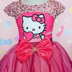 May include: A pink dress featuring a Hello Kitty graphic holding a heart. The bodice has a sequined neckline and sleeves. A large red bow with a gold clasp accents the waist. The skirt is layered with tulle.