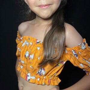 Może przedstawiać: Krótki top bez ramiączek z krótkimi rękawami w jaskrawym kolorze pomarańczowym. Top ma nadruk z kreskówkowymi psami w różnych pozach. Tkanina ma falbankę wokół dekoltu i rękawów.