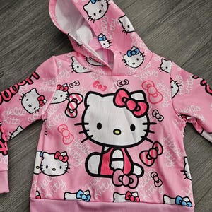 Op de afbeelding: Roze hoodie met een Hello Kitty-ontwerp. De voorkant heeft een grote Hello Kitty-afbeelding, omringd door kleinere gezichten en strikken. De tekst "Hello Kitty" herhaalt zich. Het heeft een capuchon met trekkoord en geribde manchetten.