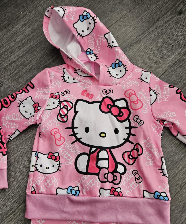 Op de afbeelding: Roze hoodie met een Hello Kitty-ontwerp. De voorkant heeft een grote Hello Kitty-afbeelding, omringd door kleinere gezichten en strikken. De tekst "Hello Kitty" herhaalt zich. Het heeft een capuchon met trekkoord en geribde manchetten.