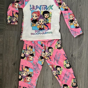 May include: Pink pajama set with a white long-sleeved top and matching pants. The top features a graphic of cartoon characters with the text "HUNTRAX K-POP DEMON HUNTERS". The pants and sleeves are covered in a repeating pattern of the same characters.