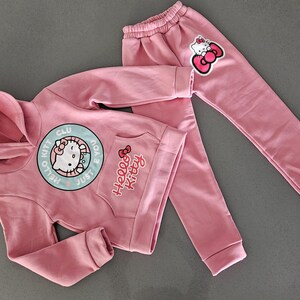 K&ouml;nnte beinhalten: Ein rosa Hello Kitty Hoodie und ein passendes Jogginghosen-Set. Der Hoodie hat eine gro&szlig;e runde Grafik mit Hello Kitty und dem Text "Just Hello Kitty Club". Die Jogginghose hat einen Hello Kitty-Aufn&auml;her am Bein.