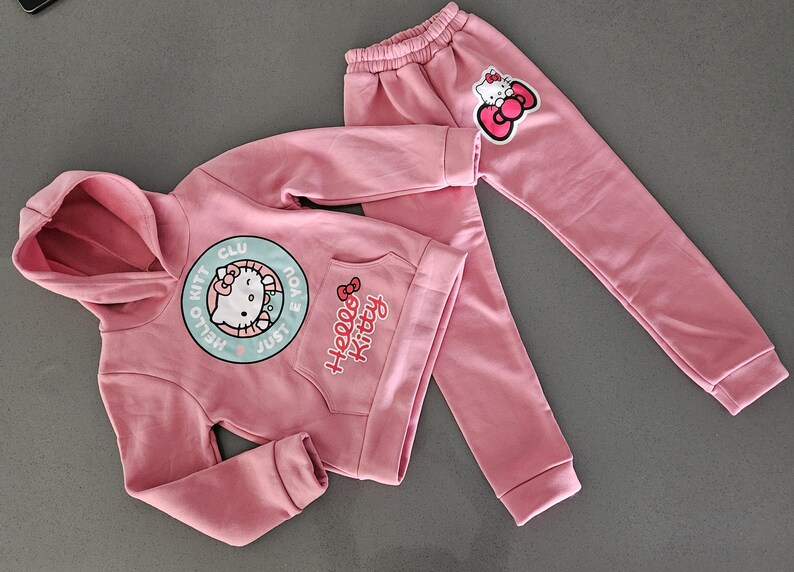 K&ouml;nnte beinhalten: Ein rosa Hello Kitty Hoodie und ein passendes Jogginghosen-Set. Der Hoodie hat eine gro&szlig;e runde Grafik mit Hello Kitty und dem Text "Just Hello Kitty Club". Die Jogginghose hat einen Hello Kitty-Aufn&auml;her am Bein.