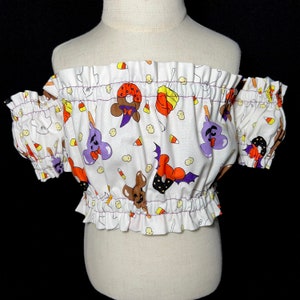 Może przedstawiać: Biały top bez ramiączek z nadrukiem o tematyce Halloween, przedstawiającym kukurydzę cukrową, duchy, nietoperze i inne straszne postacie. Top ma falbankę i jest wykonany z miękkiej, lekkiej tkaniny.