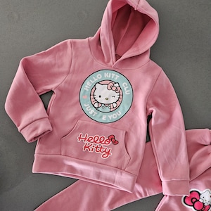 K&ouml;nnte beinhalten: Rosa Hello Kitty Hoodie und Jogginghose. Der Hoodie hat eine Vordertasche und ein rundes Grafikdesign mit Hello Kitty und dem Text "Hello Kitt Club Just E You". Die Jogginghose hat eine Hello Kitty Grafik.