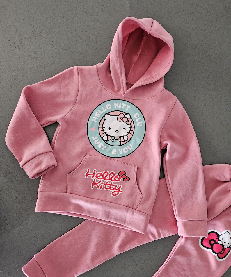 K&ouml;nnte beinhalten: Rosa Hello Kitty Hoodie und Jogginghose. Der Hoodie hat eine Vordertasche und ein rundes Grafikdesign mit Hello Kitty und dem Text "Hello Kitt Club Just E You". Die Jogginghose hat eine Hello Kitty Grafik.