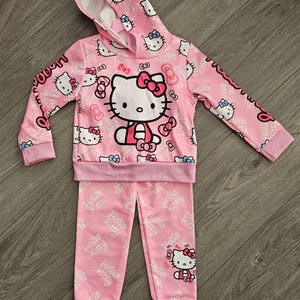 Op de afbeelding: Roze Hello Kitty-set bestaande uit een hoodie en een bijpassende broek. De hoodie heeft een grote Hello Kitty-afbeelding en een herhalend patroon van Hello Kitty en strikken. Op de mouwen en broek staat "Hello Kitty".