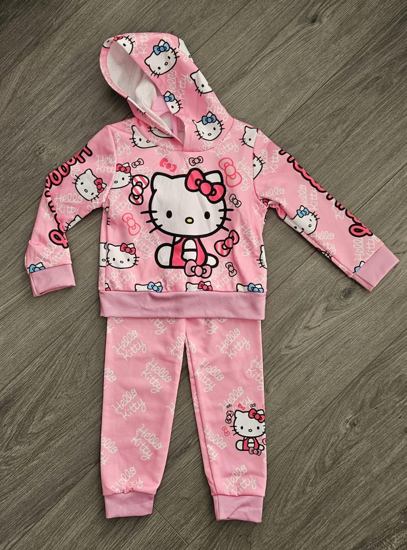 Op de afbeelding: Roze Hello Kitty-set bestaande uit een hoodie en een bijpassende broek. De hoodie heeft een grote Hello Kitty-afbeelding en een herhalend patroon van Hello Kitty en strikken. Op de mouwen en broek staat "Hello Kitty".