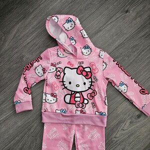 Op de afbeelding: Roze Hello Kitty hoodie en bijpassende broek. De set heeft een herhalend patroon van Hello Kitty gezichten en strikken. De hoodie heeft een grote Hello Kitty afbeelding op de voorkant. De tekst "Hello Kitty" is gedrukt.