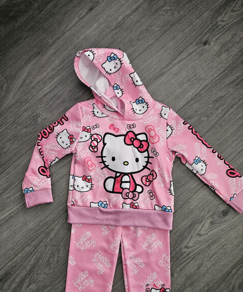 Op de afbeelding: Roze Hello Kitty hoodie en bijpassende broek. De set heeft een herhalend patroon van Hello Kitty gezichten en strikken. De hoodie heeft een grote Hello Kitty afbeelding op de voorkant. De tekst "Hello Kitty" is gedrukt.