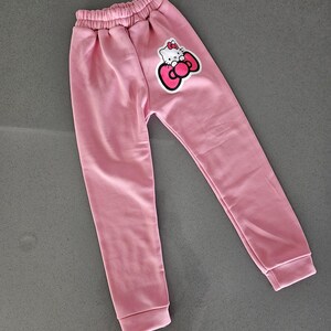 K&ouml;nnte beinhalten: Hellrosa Jogginghose mit elastischem Bund und B&uuml;ndchen. Auf dem linken Bein befindet sich eine Hello Kitty-Grafik mit roter Schleife. Die Hose ist aus weichem Material.