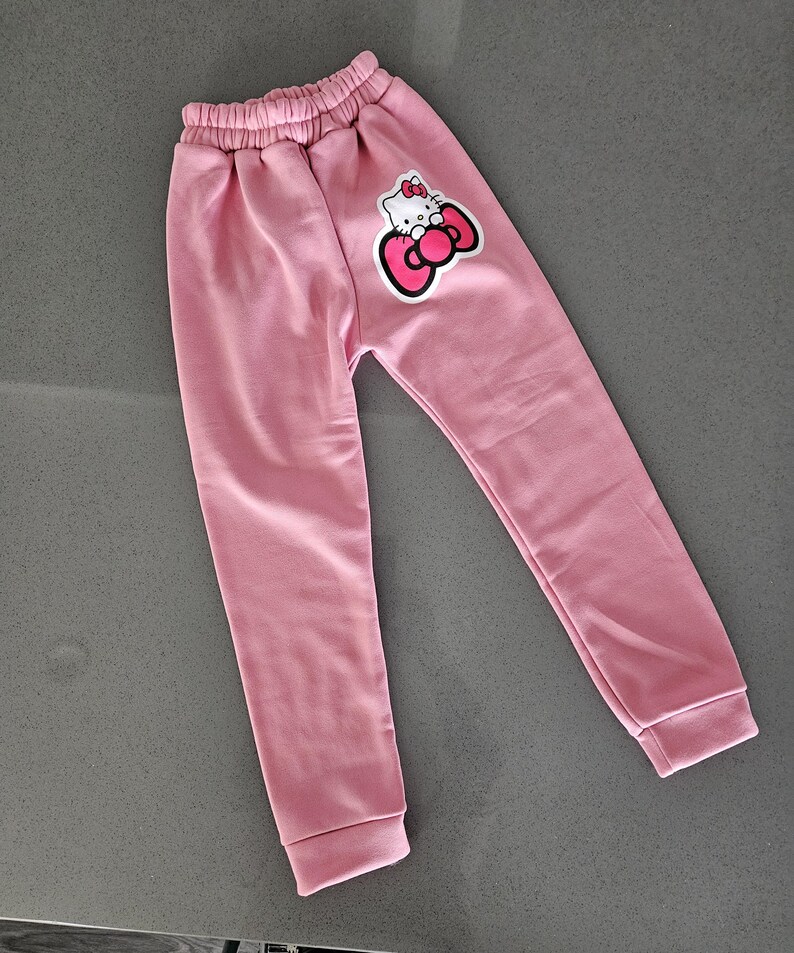 K&ouml;nnte beinhalten: Hellrosa Jogginghose mit elastischem Bund und B&uuml;ndchen. Auf dem linken Bein befindet sich eine Hello Kitty-Grafik mit roter Schleife. Die Hose ist aus weichem Material.
