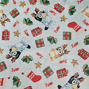 Pode incluir: Tecido azul claro com um padrão de Natal repetido. O design inclui cães de desenho animado com chifres de rena, presentes embrulhados, bengalas doces, azevinho, estrelas e meias de Natal vermelhas.
