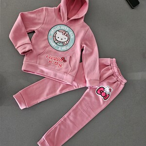 K&ouml;nnte beinhalten: Ein rosa Hello Kitty-Set, bestehend aus einem Kapuzenpullover und einer passenden Jogginghose. Der Kapuzenpullover hat eine Vordertasche und ein Hello Kitty-Motiv. Die Hose hat einen elastischen Bund und ein Hello Kitty-Motiv am Bein.