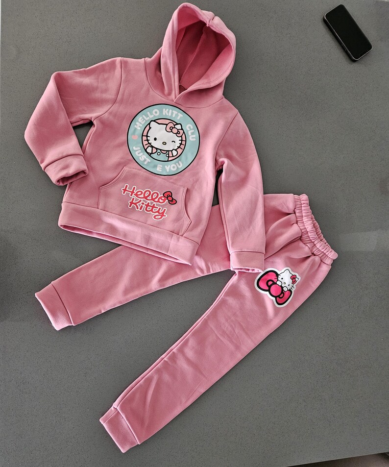 K&ouml;nnte beinhalten: Ein rosa Hello Kitty-Set, bestehend aus einem Kapuzenpullover und einer passenden Jogginghose. Der Kapuzenpullover hat eine Vordertasche und ein Hello Kitty-Motiv. Die Hose hat einen elastischen Bund und ein Hello Kitty-Motiv am Bein.