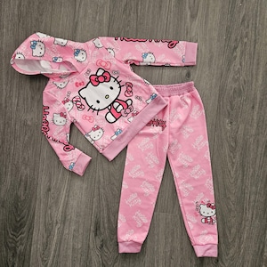 Op de afbeelding: Roze Hello Kitty hoodie en bijpassende joggingbroek set. De hoodie heeft een grote Hello Kitty afbeelding en kleinere prints. De joggingbroek heeft een all-over print van het Hello Kitty logo. De set is gemaakt van een zacht materiaal.