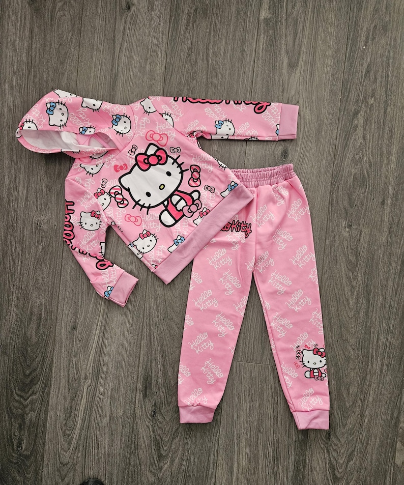 Op de afbeelding: Roze Hello Kitty hoodie en bijpassende joggingbroek set. De hoodie heeft een grote Hello Kitty afbeelding en kleinere prints. De joggingbroek heeft een all-over print van het Hello Kitty logo. De set is gemaakt van een zacht materiaal.