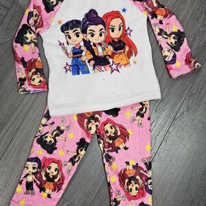 May include: Pink and white pajama set featuring a cartoon graphic. The long-sleeved top has a white body with pink sleeves and a pink neckline. The sleeves and pants are covered in a repeating cartoon character pattern.