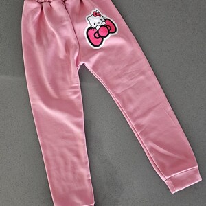 K&ouml;nnte beinhalten: Hellrosa Jogginghose mit elastischem Bund und gerippten B&uuml;ndchen. Auf dem linken Bein befindet sich eine Cartoon-Grafik einer Katze mit Schleife. Die Hose ist aus weichem Material.
