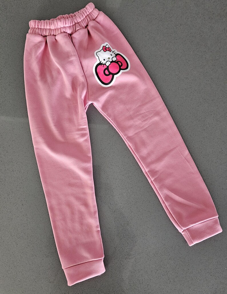 K&ouml;nnte beinhalten: Hellrosa Jogginghose mit elastischem Bund und gerippten B&uuml;ndchen. Auf dem linken Bein befindet sich eine Cartoon-Grafik einer Katze mit Schleife. Die Hose ist aus weichem Material.
