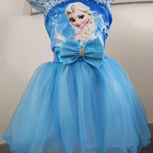 Op de afbeelding: Een lichtblauwe prinsessenjurk met een tule rok en een lijfje met een cartoonafbeelding van een prinses. De jurk heeft een grote strik in de taille en is versierd met pailletten rond de halslijn.