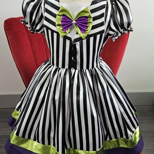 May include: A black and white striped dress with a purple and green bow. The dress has short, puffed sleeves and a full skirt with purple and green trim. The dress is displayed on a mannequin.