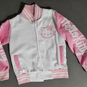 Op de afbeelding: Een wit en roze Hello Kitty varsity jack. De jas heeft roze mouwen met witte "Hello Kitty" letters, een roze kraag en een Hello Kitty afbeelding op de borst. Het heeft een knoopsluiting en gestreepte manchetten.
