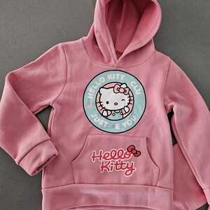 K&ouml;nnte beinhalten: Rosa Hello Kitty Hoodie und passende Hose. Der Hoodie hat eine Vordertasche und eine runde Grafik mit Hello Kitty und dem Text "Hello Kitt Club Just E You". Das Hello Kitty-Logo befindet sich ebenfalls auf der Vordertasche.
